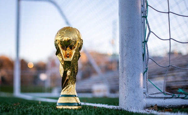 Close up photo of the World Cup trophy on a soccer field with a goal out of focus in the background.